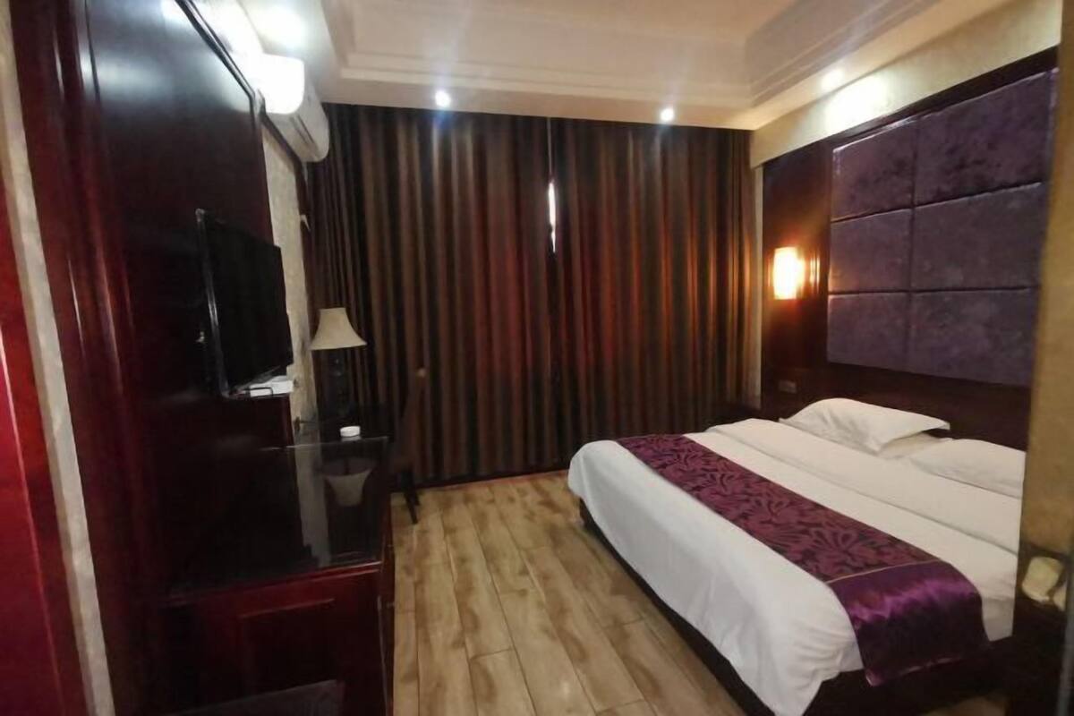 A hotel room with a wooden floor, a bed with white bedding and a purple throw, a TV on a wooden cabinet, and a wall-mounted light fixture.