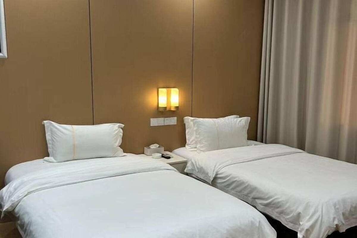 A hotel room with two single beds, a nightstand with a phone and a lamp, and a wall-mounted light fixture.