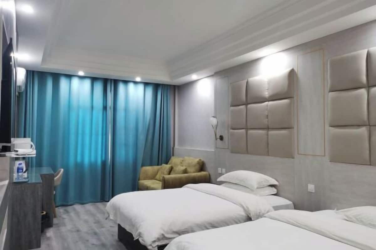 A hotel room with two beds, a desk with a chair, a sofa, and a large window with blue curtains.