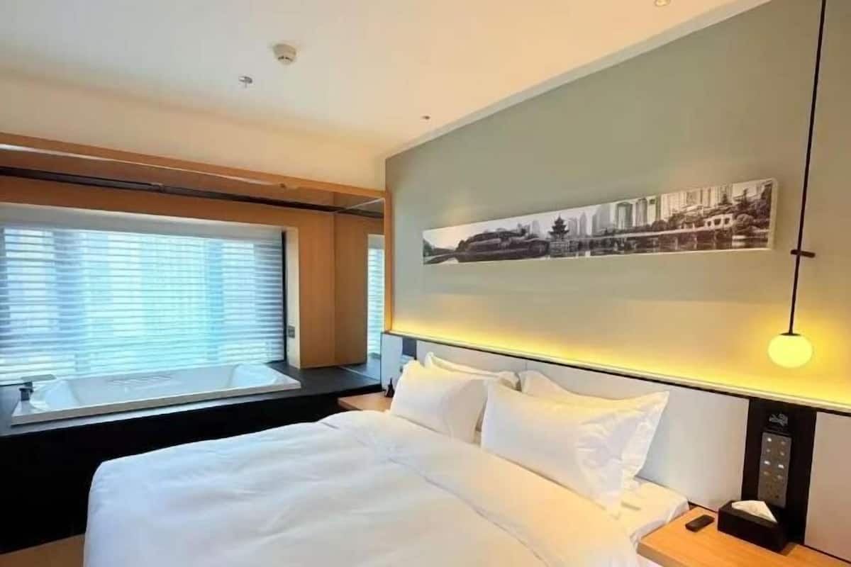 A hotel room with a bed, a window with blinds, a wall-mounted picture, and a bedside table with a lamp.