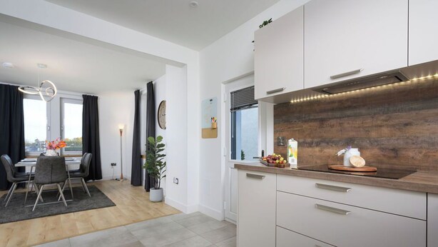 A modern kitchen with white cabinets, a wooden countertop, and a built-in stove. There is a dining area with a table and chairs, and a potted plant.