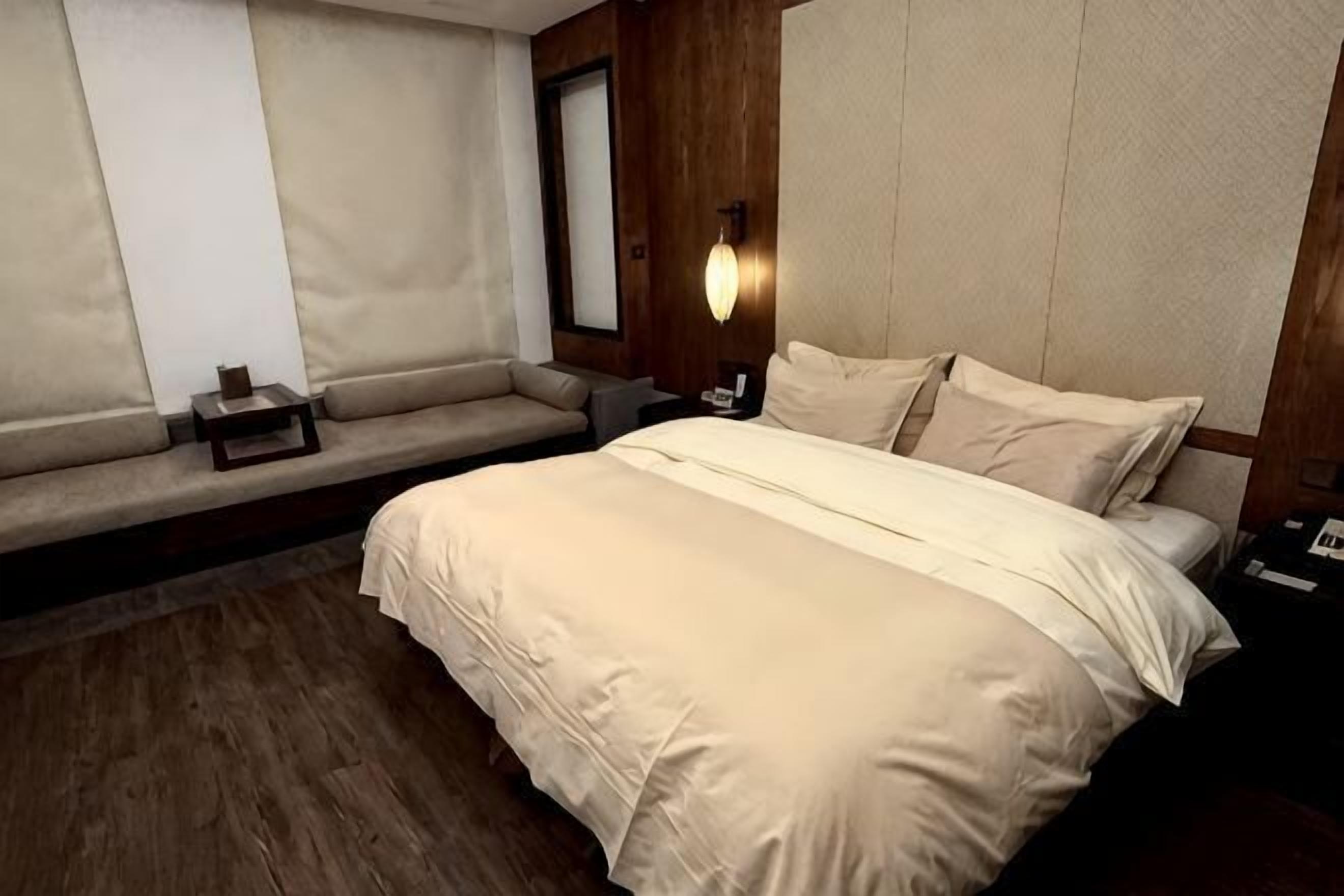 A hotel room with a bed, a bench, a nightstand, and a wall-mounted light fixture.