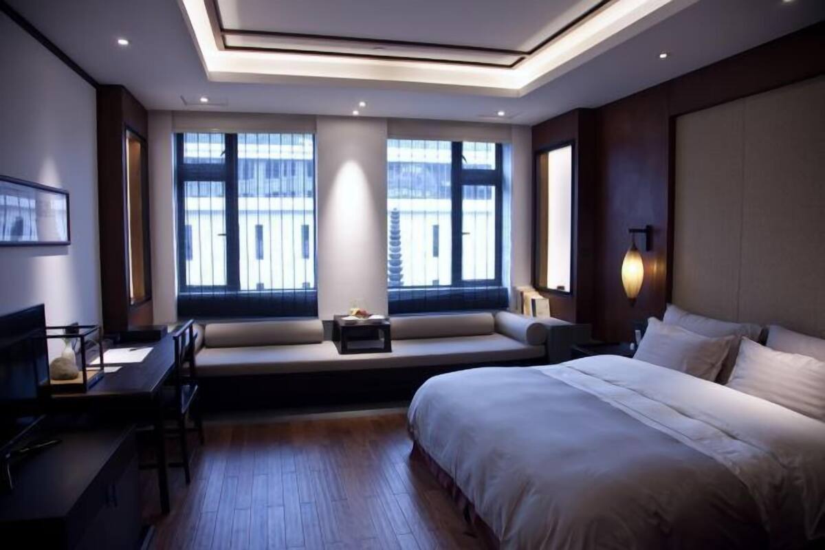 A modern hotel room with a large bed, a seating area with a desk, and large windows offering a city view.