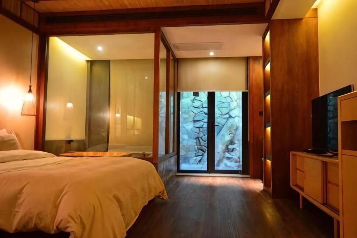 A bedroom with a bed, a TV, a wooden cabinet, and a large window with a view of trees.