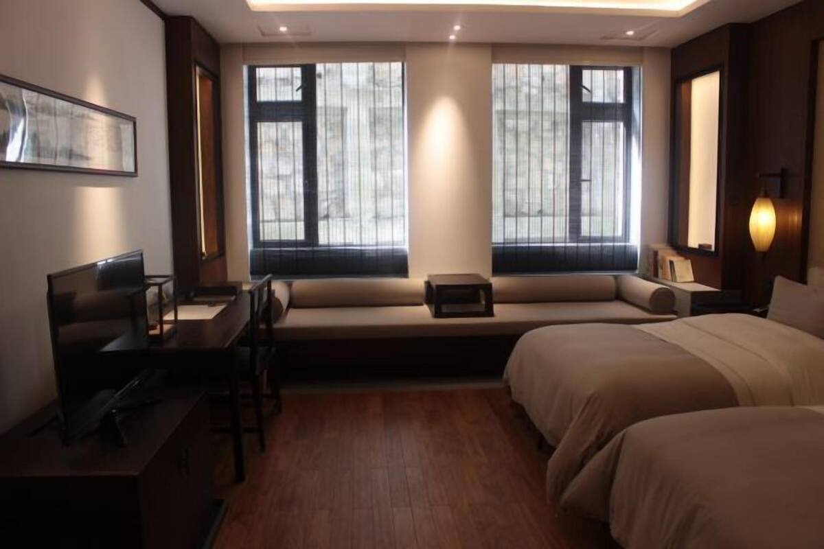 A hotel room with a desk, two beds, a sofa, and large windows.