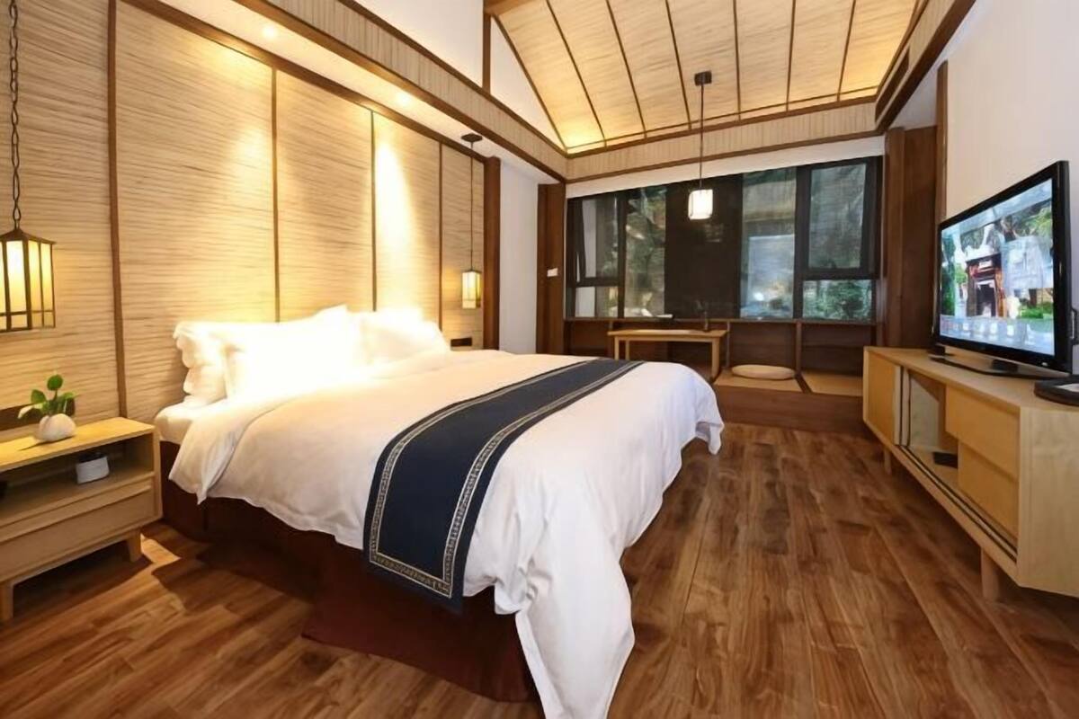 A modern hotel room with a large bed, wooden flooring, a television, and a view of the outdoors through large windows.