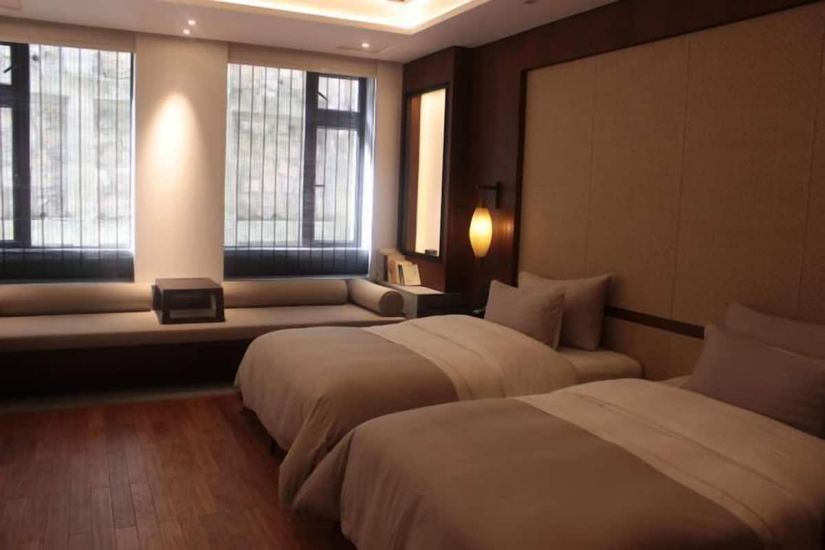 A hotel room with two beds, a bench with a small table, and a large window with curtains.