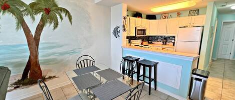 A kitchen with white cabinets, a microwave, a refrigerator, and a coffee maker. There is a dining table with chairs and a mural of a palm tree on the wall.