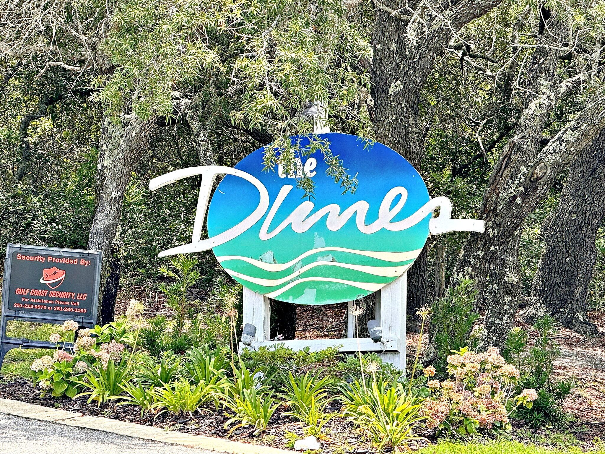 A sign for 'The Dunes' with a beach theme, surrounded by greenery and a security sign.
