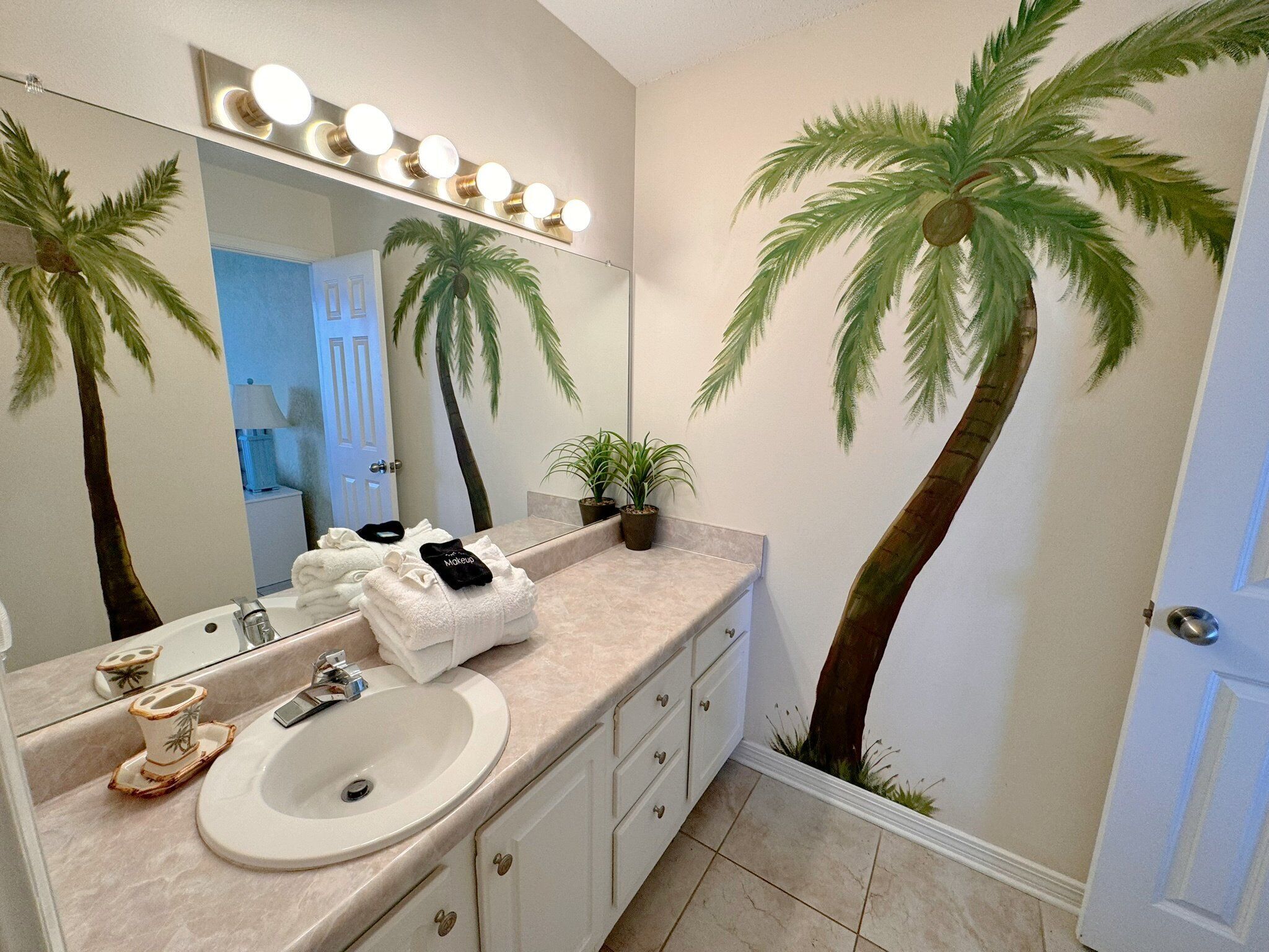 A bathroom with a vanity, a sink, a mirror with lights, and palm tree wall art.