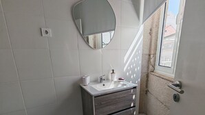 Une salle de bain avec des carreaux blancs, un meuble-lavabo en bois, un miroir rond et une fenêtre donnant sur des immeubles.