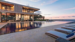Um resort moderno com uma grande piscina infinita, espreguiçadeiras e vista para o oceano ao pôr do sol.
