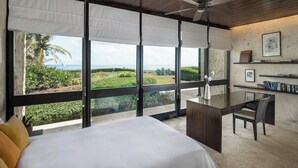 Quarto com cama grande, escrivaninha e cadeira, com vista para uma paisagem exuberante e o oceano.