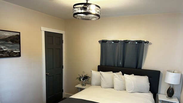 A neatly arranged bedroom with a bed, a nightstand, a lamp, a door, and a wall-mounted picture. - This cozy house has been completely remodeled inside and features three bedrooms. (Helena)
