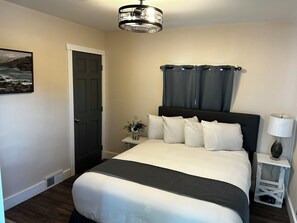 A neatly arranged bedroom with a bed, a nightstand, a lamp, a door, and a wall-mounted picture. - This cozy house has been completely remodeled inside and features three bedrooms. (Helena)