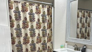 A bathroom with a shower curtain featuring a leaf pattern, a sink with a mirror above it, and dark cabinets. - This cozy house has been completely remodeled inside and features three bedrooms. (Helena)