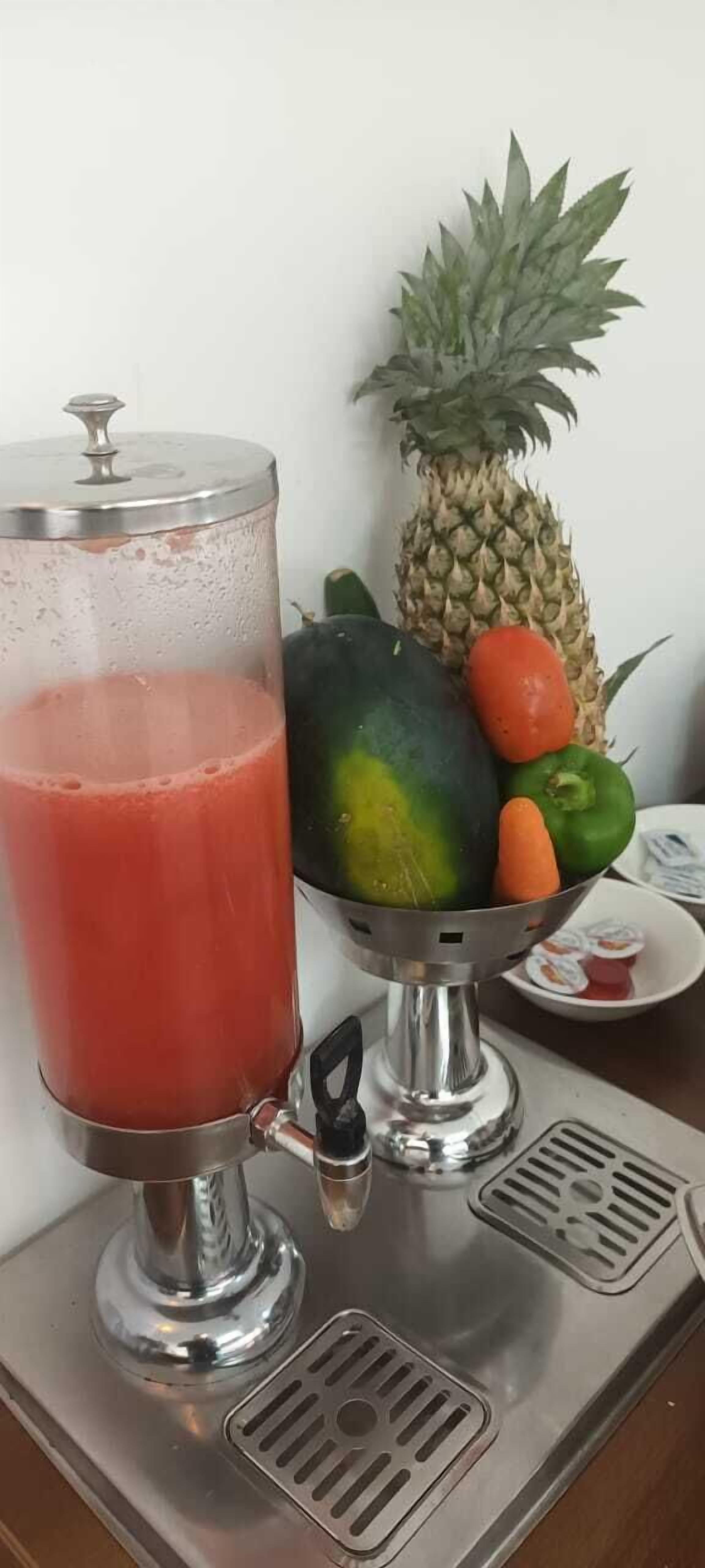 Ein Getränkespender mit einem Glas rotem Getränk und einer Schüssel Obst, darunter Ananas, Melone und Paprika.
