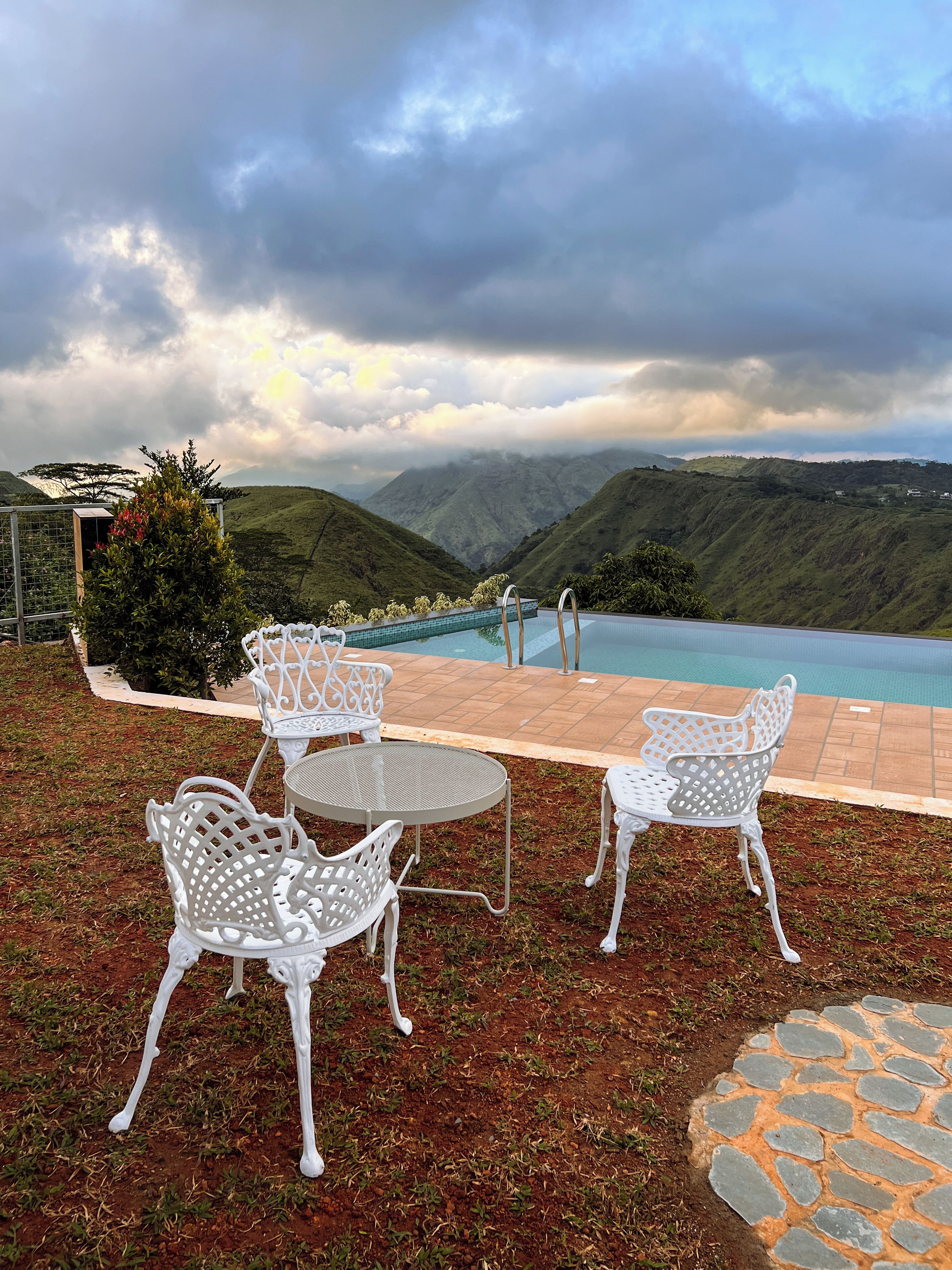 Zona junto a la piscina con sillas y una mesa de metal blanco, con vistas a un paisaje montañoso.