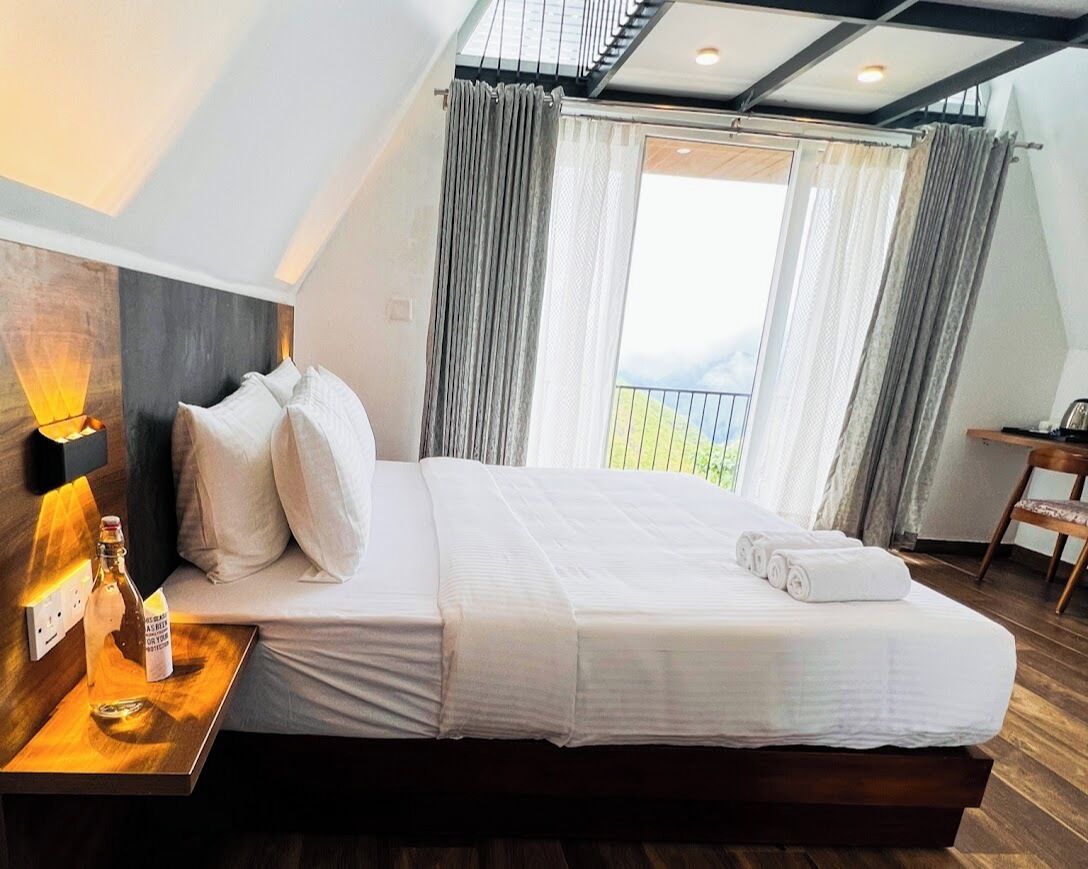 A hotel room with a large bed, a bedside table with a bottle, and a view of the outdoors through a window.