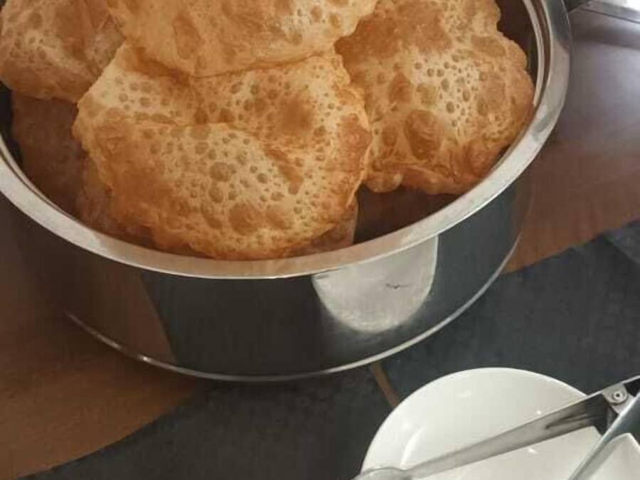 Un cuenco metálico lleno de panes planos dorados y esponjosos, junto a un plato blanco y un par de pinzas sobre un paño oscuro.