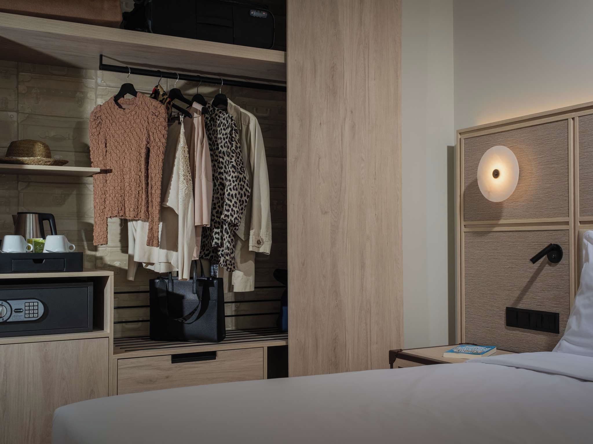 Habitación de hotel con cama, mesita de noche con lámpara, armario con ropa, cafetera y radio.