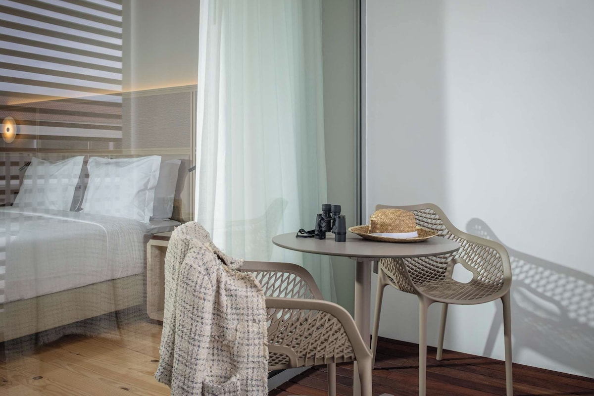 Ein modernes Hotelzimmer mit einem Bett, einem kleinen Tisch mit Fernglas und einem Korbstuhl.