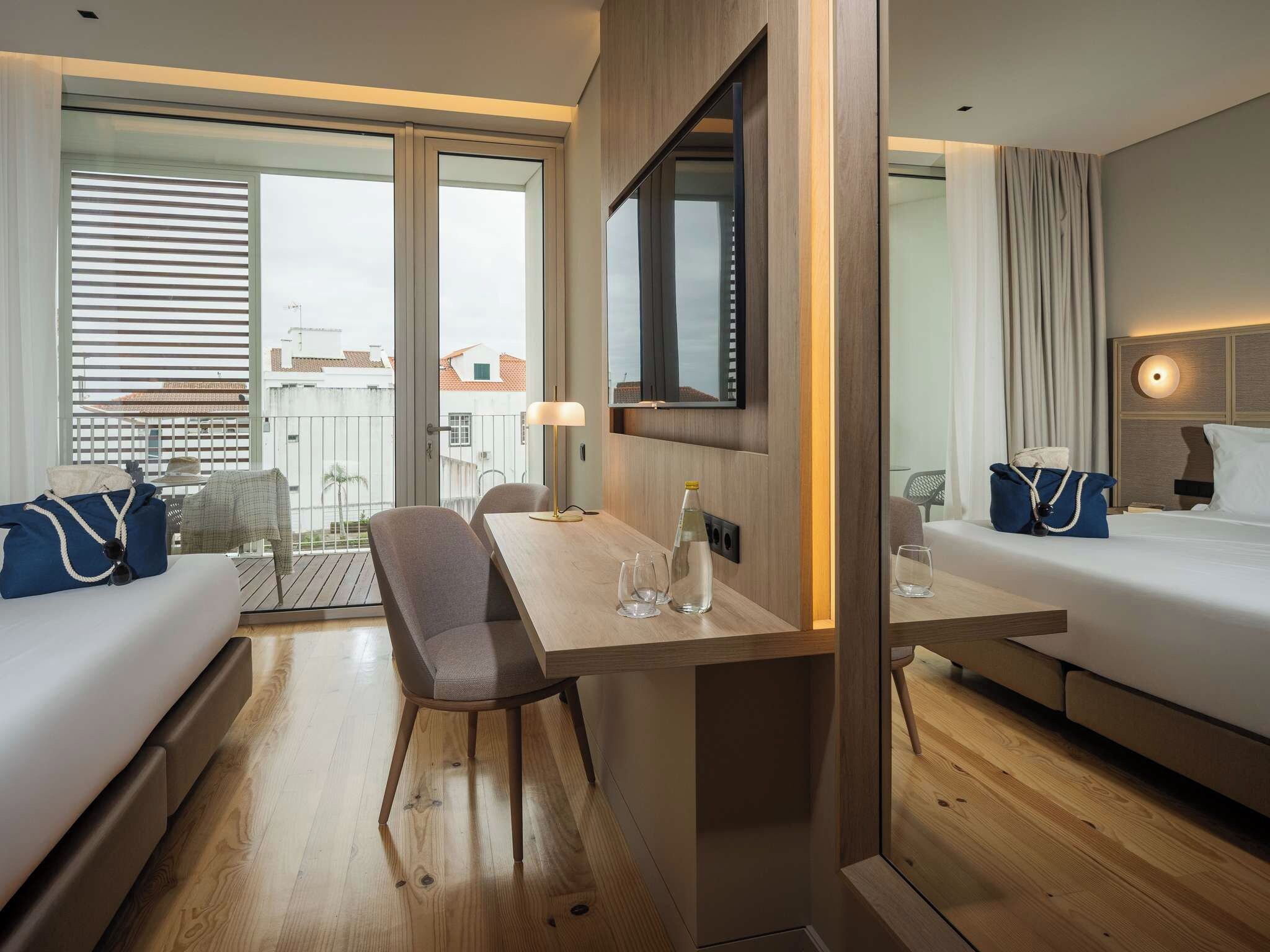 Habitación de hotel con suelo de madera, una mesa de comedor con sillas, un sofá, una cama, un televisor y un balcón con vistas a edificios.