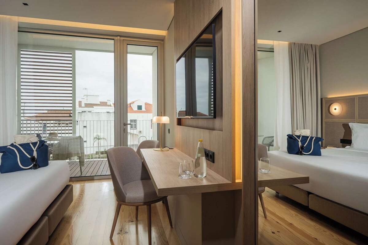Ein Hotelzimmer mit Holzboden, einem Esstisch mit Stühlen, einer Couch, einem Bett, einem Fernseher und einem Balkon mit Blick auf Gebäude.