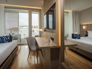 Ein Hotelzimmer mit Holzboden, einem Esstisch mit Stühlen, einer Couch, einem Bett, einem Fernseher und einem Balkon mit Blick auf Gebäude.