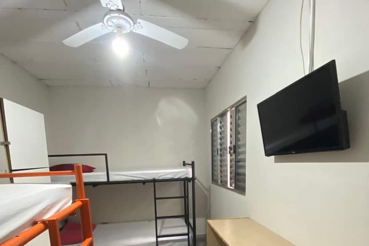 A dormitory room with bunk beds, a ceiling fan, a TV mounted on the wall, and a wooden bench.