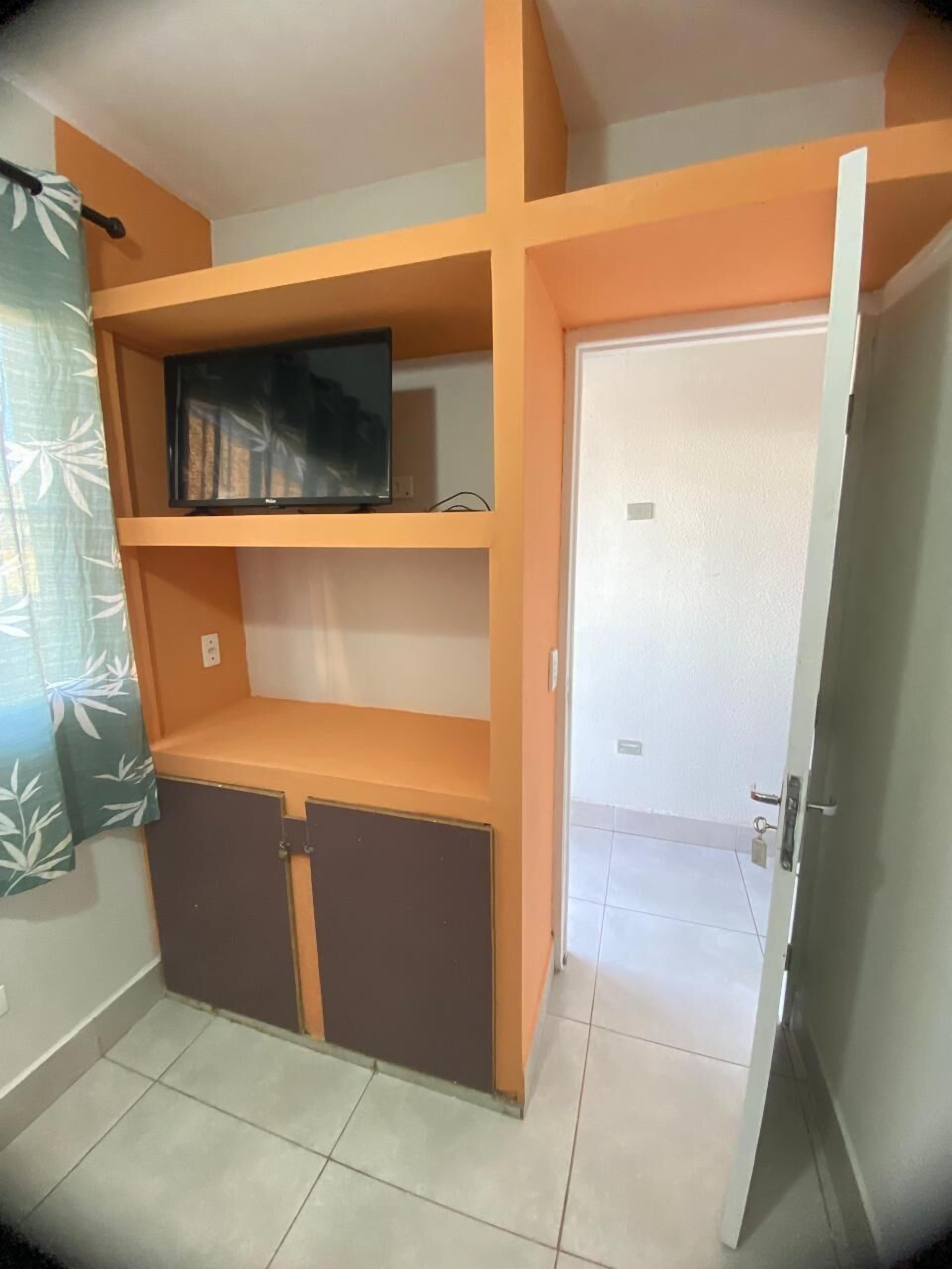 Quarto com prateleira para televisão, guarda-roupa e porta que dá para outro espaço.