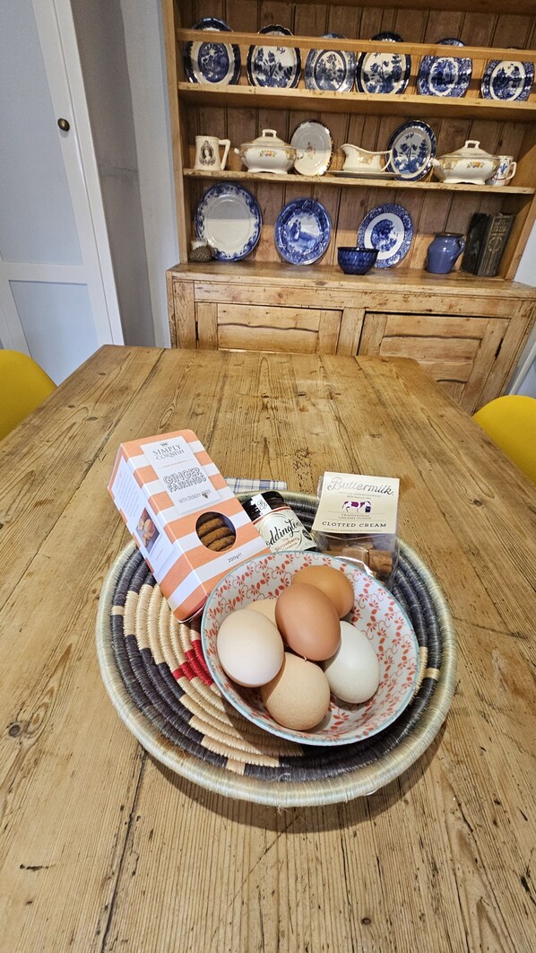 A wooden table with a woven placemat holding a bowl of eggs and a box of ginger biscuits. Behind the table, there is a shelf with blue and white ceramic dishes.