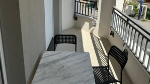 Un balcón con una mesa de mármol y dos sillas, una barandilla pequeña, y vistas a edificios cercanos.