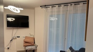 Una sala de estar moderna con una televisión de pantalla plana, un tapete rojo, un sofá oscuro con cojines estampados y una ventana con persianas.