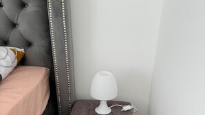 Un dormitorio con cabecera capitonada, una mesita de noche con lámpara blanca y un reposapiés gris.