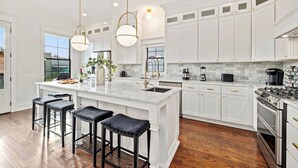 Une cuisine moderne avec des meubles blancs, un grand îlot central doté de tabourets, des appareils en acier inoxydable et un éclairage suspendu.