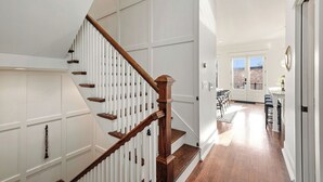 Un escalier aux panneaux blancs et aux marches en bois mène à une pièce comprenant un coin repas et offrant une vue sur l’extérieur.