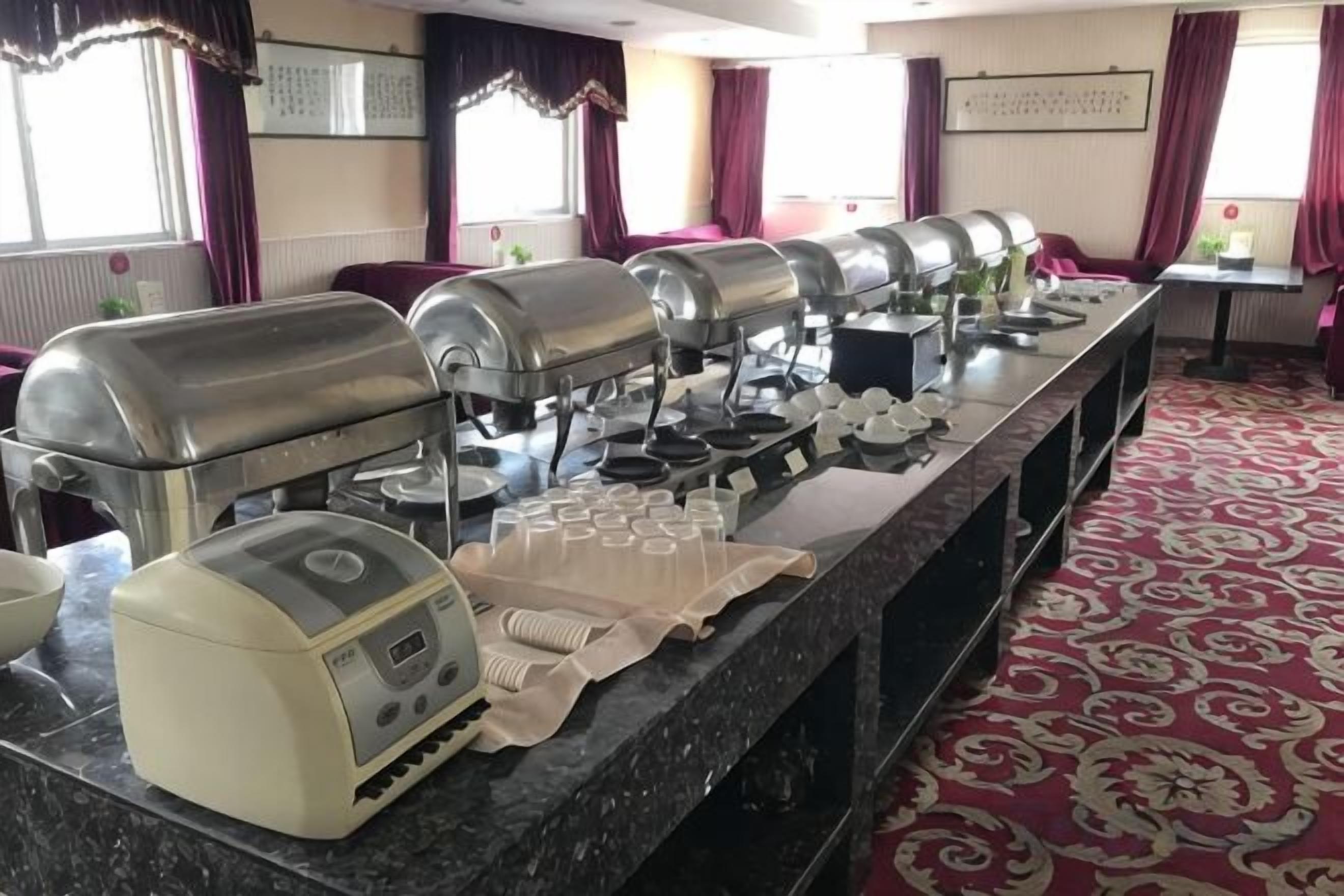 A buffet setup with multiple self-service stations, including hot food and beverages, in a room with patterned carpet and large windows.
