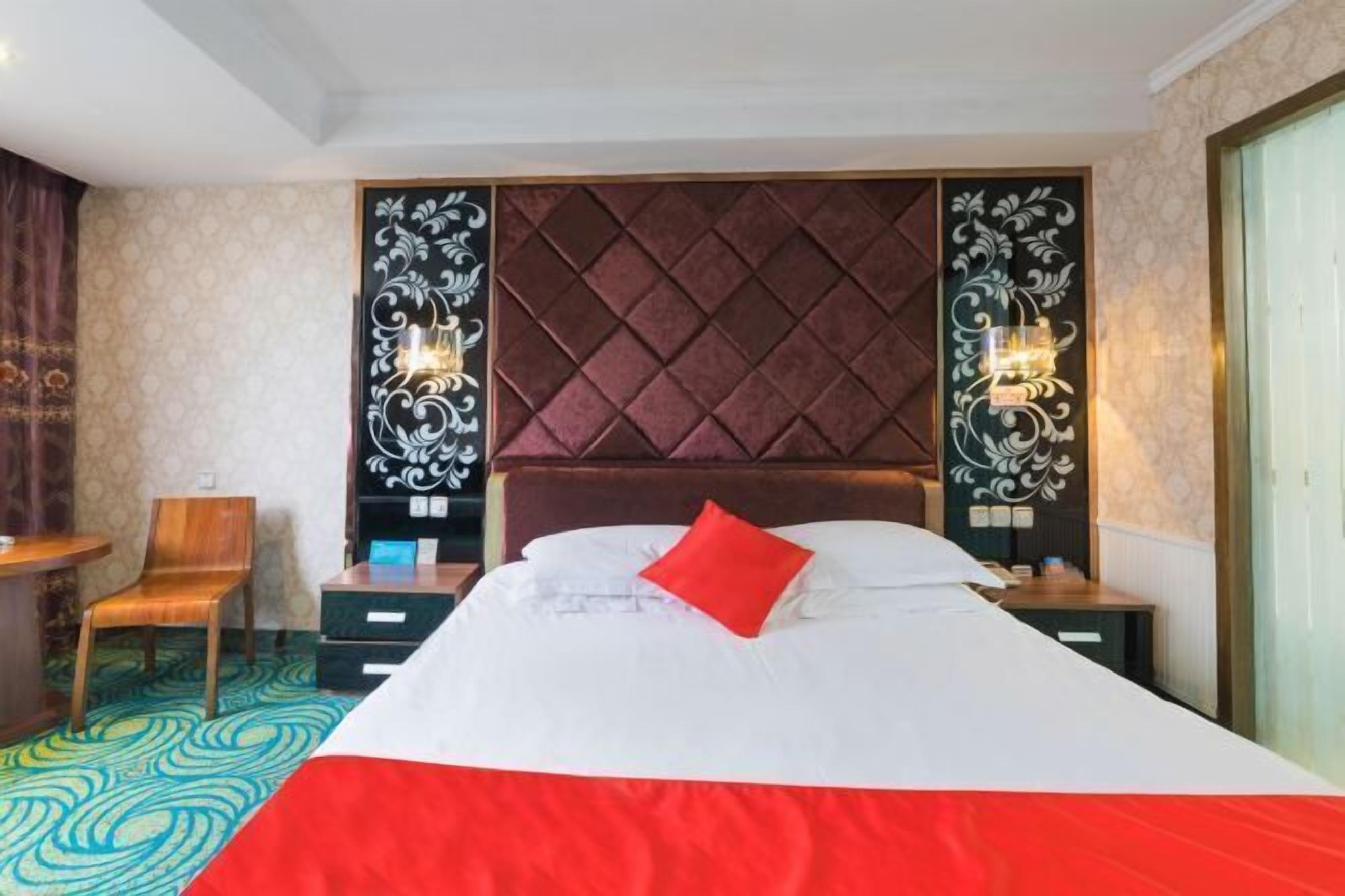 Habitación de hotel con una cama grande, cabecera de madera, dos mesitas de noche con lámparas, una silla y una alfombra estampada.