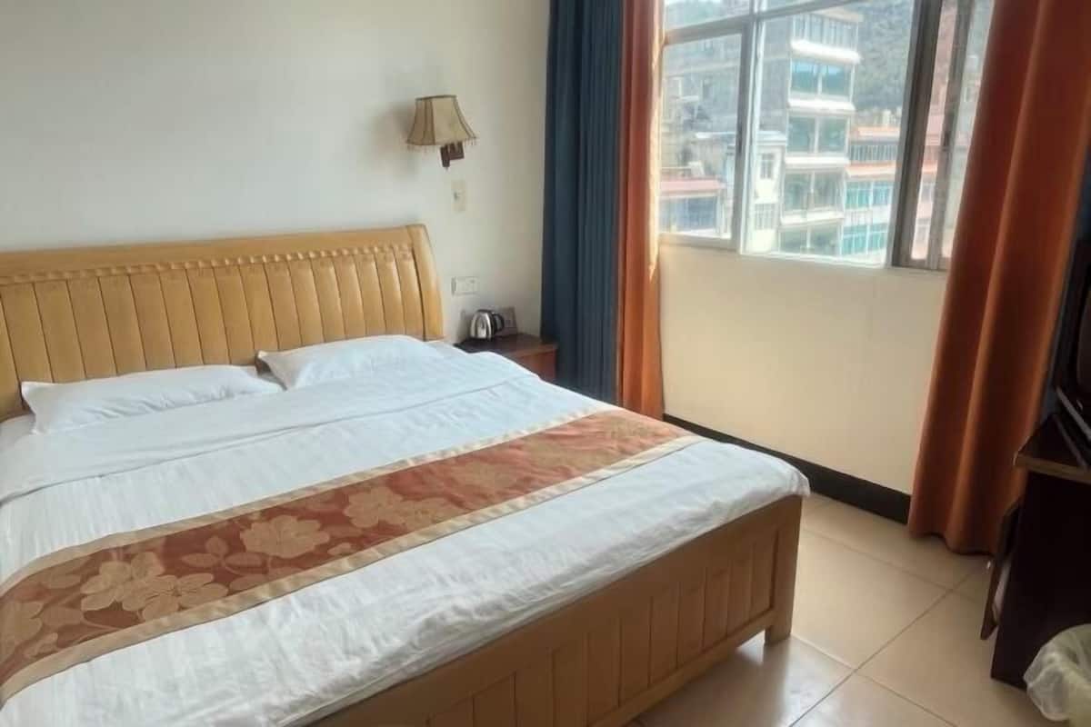 Una cama con colcha estampada, una mesita de noche con lámpara y vistas a edificios a través de una ventana.