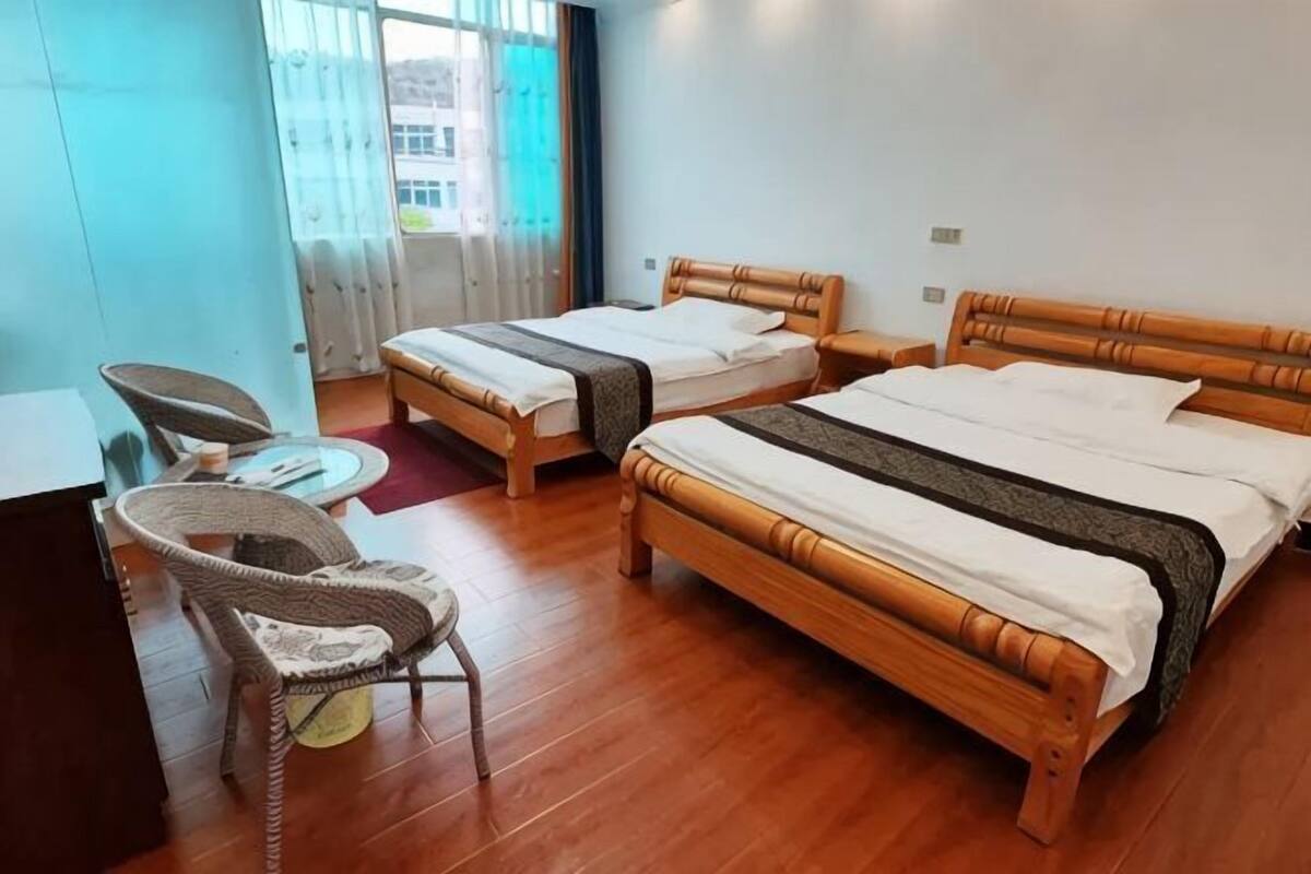 Habitación de hotel con dos camas de madera, una mesita redonda pequeña, una silla de mimbre y vistas al exterior.