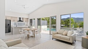 A spacious living area with a large window, a kitchen with modern appliances, and a view of a pool and mountains. - Belle Escapes Sea La Vie (Trinity Beach)