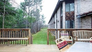 A wooden deck with a bench and chairs, a brick building with large windows, and a forested area in the background.