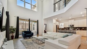 Amplia sala de estar con un sofá blanco, una mesa de centro y un televisor. Se ve una escalera que conduce a un nivel superior, y un área de cocina al fondo.