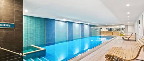 A modern indoor swimming pool with a 'No Diving' sign, a wooden bench, and a reception area in the background.
