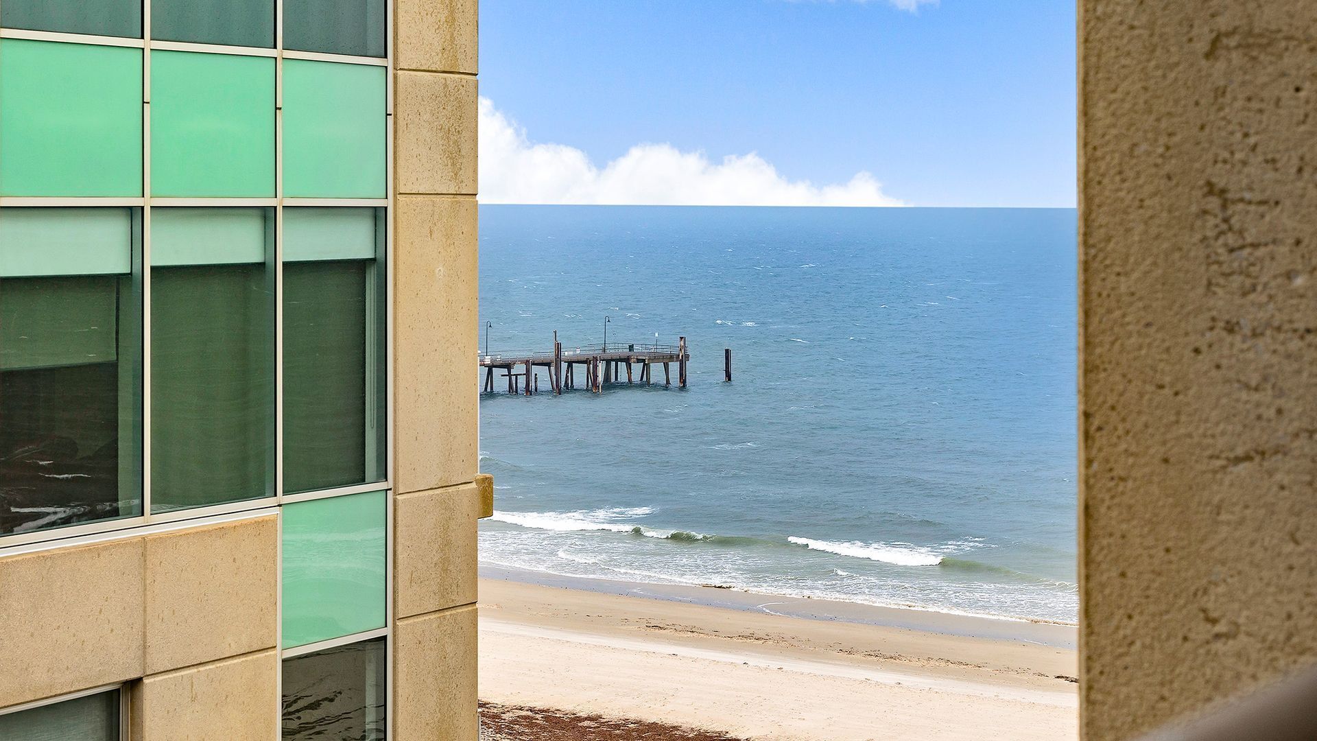 A view of a beach with a pier extending into the ocean, seen through a building with large windows.