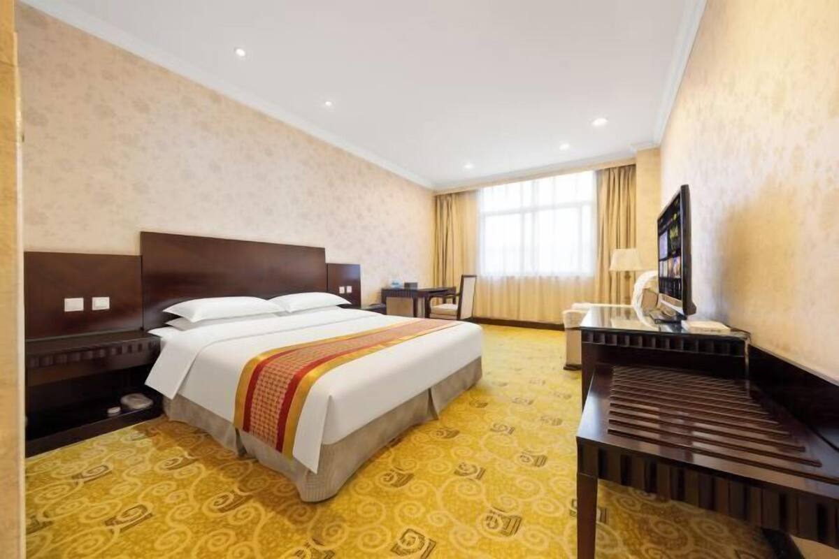 A hotel room with a bed, bedside table, desk, lamp, and a large window with curtains.