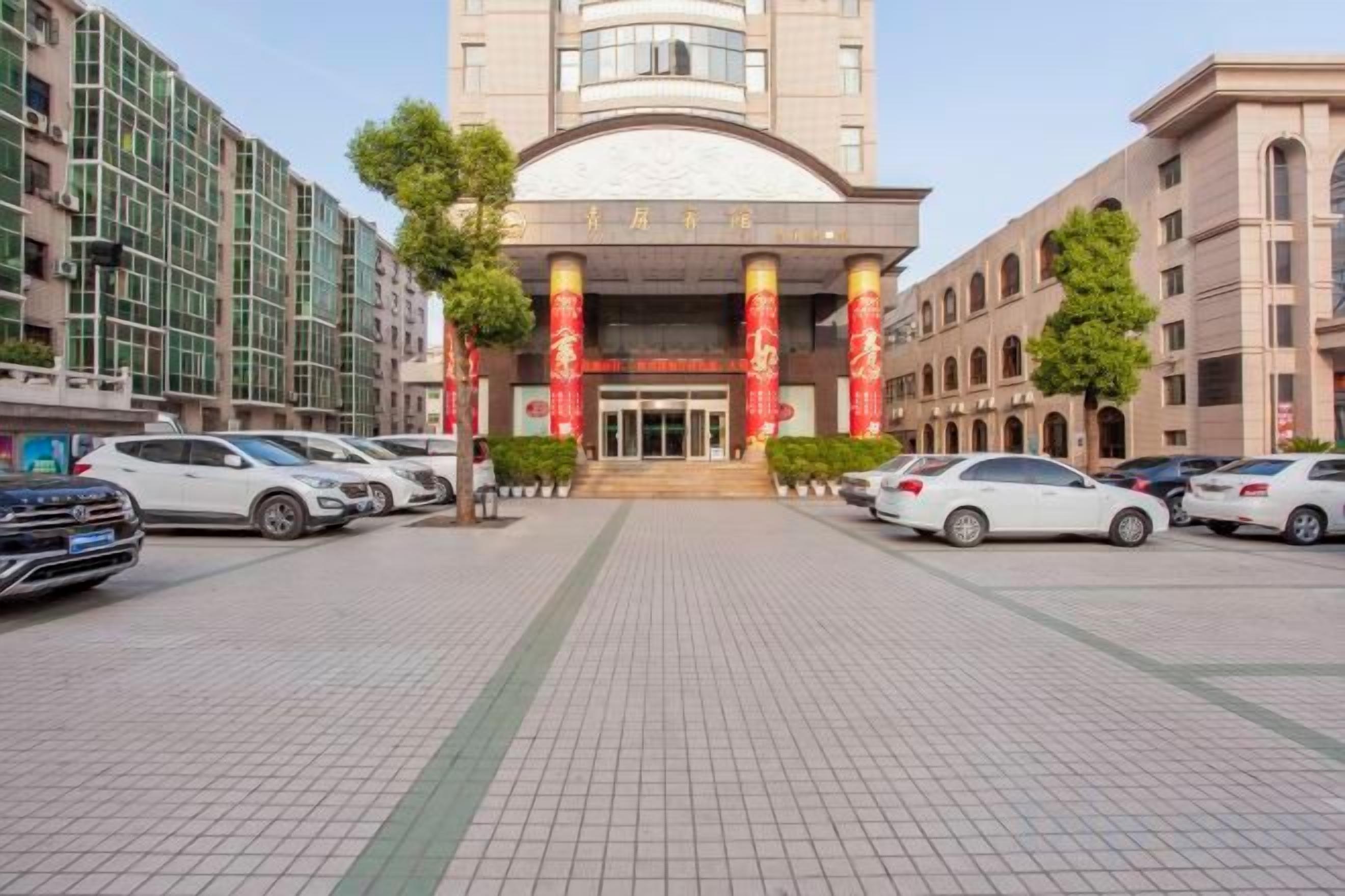 A hotel entrance with a modern facade, a parking lot, and several cars parked in front.