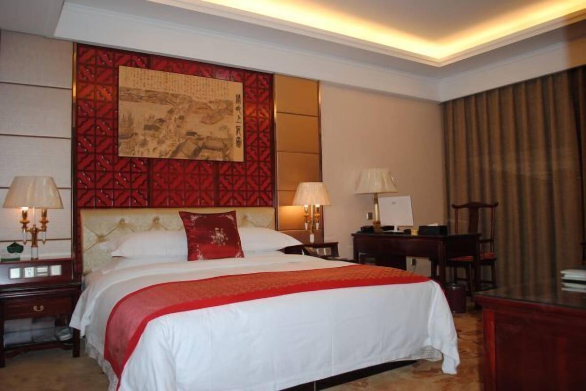 A hotel room with a bed, bedside tables, lamps, a desk, and a large framed artwork on the wall.