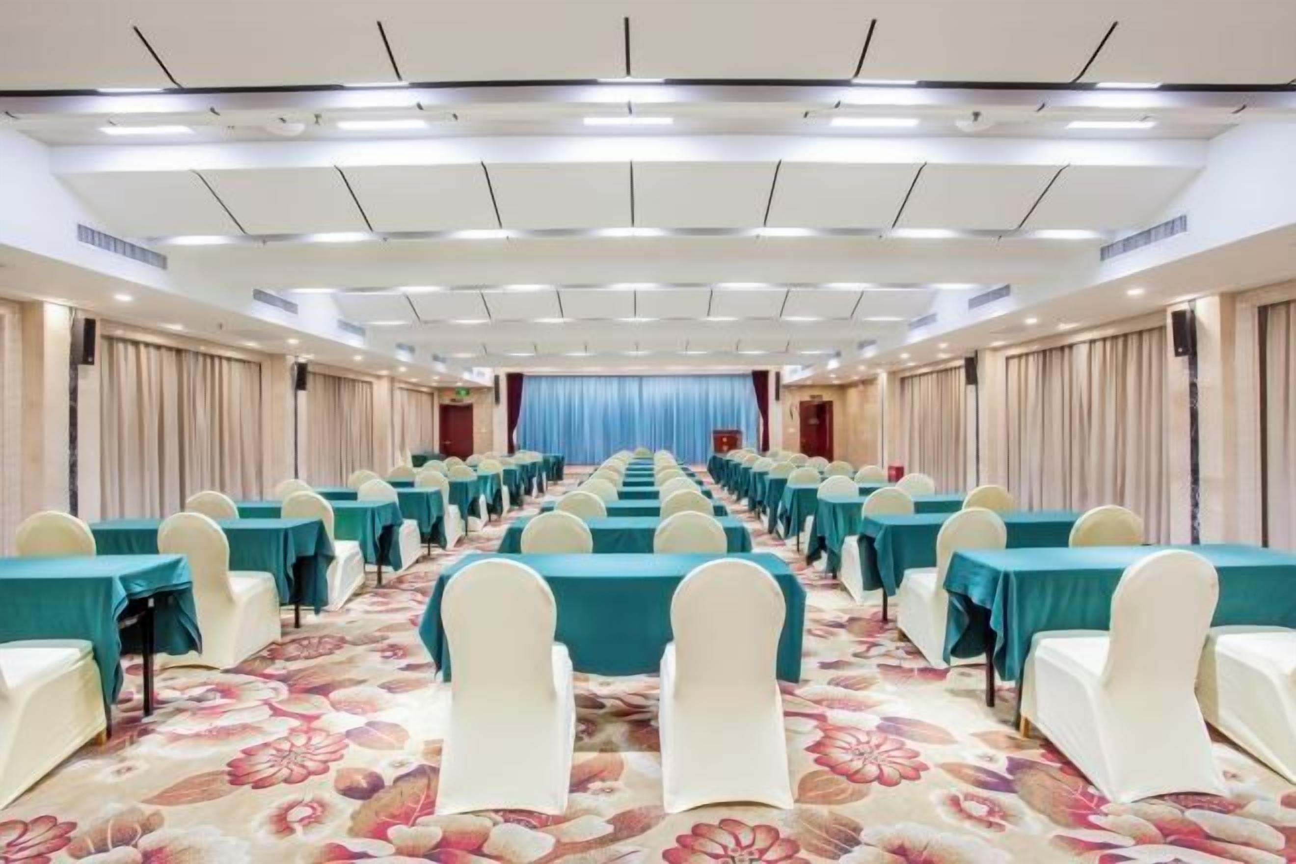 Une salle de conférence avec des rangées de tables et de chaises, une scène à l’avant et des rideaux sur les côtés.